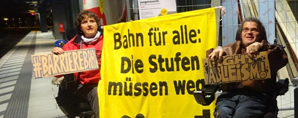 2 PAktive in Rollis mit Protestschildern am Bahngleis