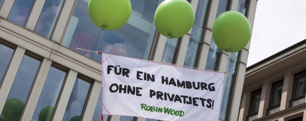 Protestbanner gegen Privatjets hängt an drei grünen Ballons in der Luft