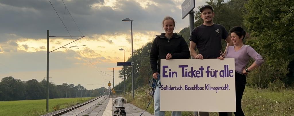 Das Angebot von Bahn und Bus in der Fläche muss ausgebaut und mit einem sozial gerechten Ticket für alle kombiniert werden (Foto: Raphael / ROBIN WOOD) Drei Wartende mit Hund und Schild "Ein Ticket für alle" auf einem Bahnsteig