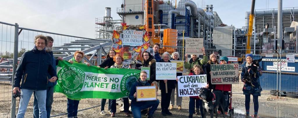 Parents for Future Cuxhaven & BUND Kreisgruppe Cuxhaven Menschen mit vielen Schildern, Bannern und brennendem PappBaum vor einem Kraftwerk
