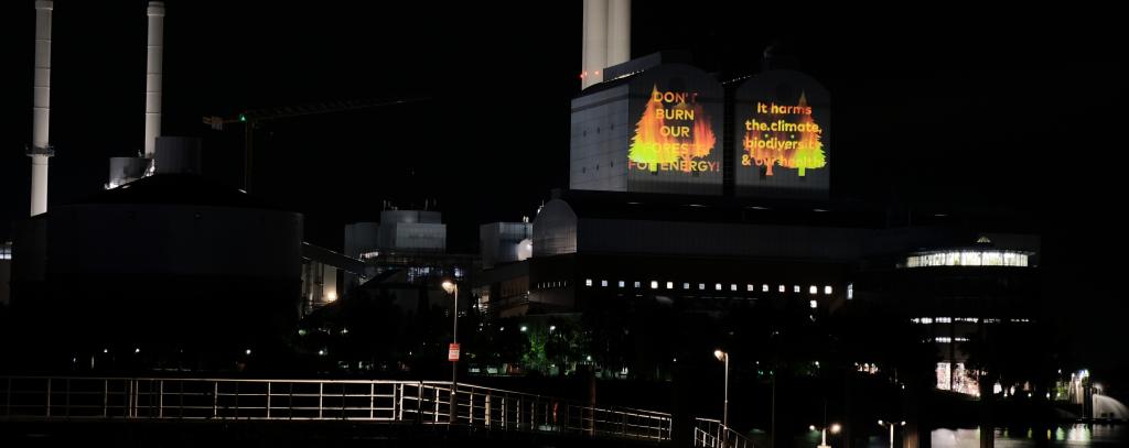Kraftwerk im Dunkeln mit projiziertem Slogan gegen Holzverbrennung