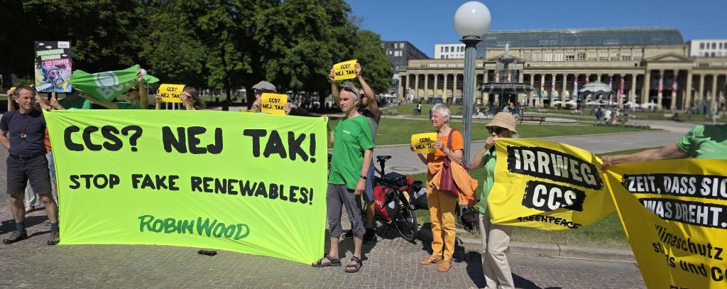Foto: ROBIN WOOD Viele Menschen mit einem Neongelben Banner und mehreren Schildern gucken entschlossen.