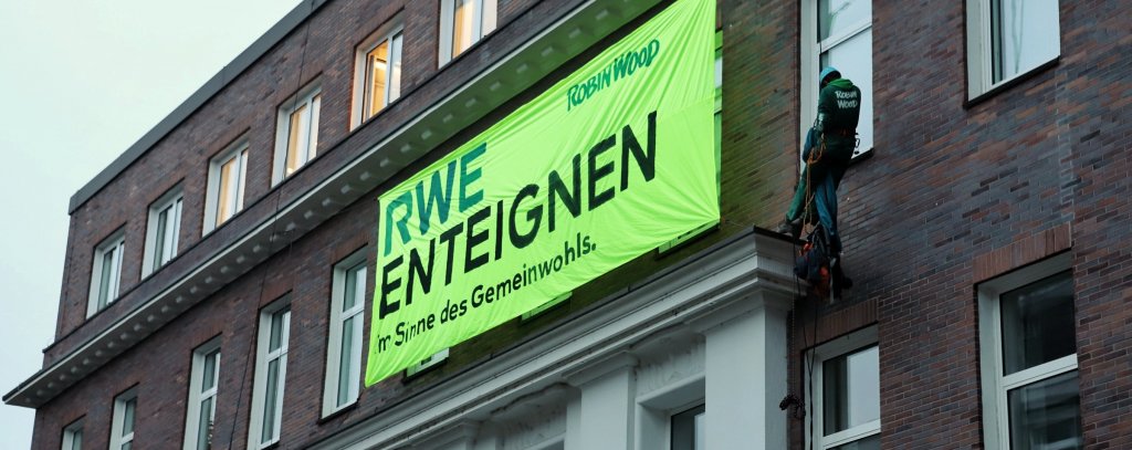 Zwei Kletter*innen haben sich mit einem Banner vom Dach der RWE-Zentrale in Essen abgeseilt. Auf dem Banner steht: RWE enteignen - Sim Sinne des Gemeinwohls