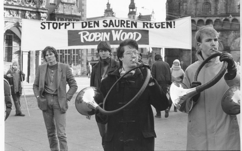 Aktivisten mit Blasinstrumenten und einem Banner "Stoppt den sauren Regen!"