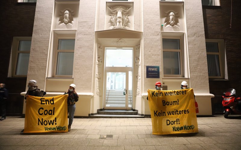 Vor der RWE-Zentrale halten weitere Aktivist*innen Banner mit den Aufschriften "End Coal Now" und "Kein weiteres Dorf. Kein weiterer Baum"