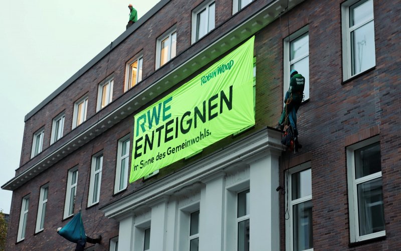 Zwei Kletter*innen haben sich mit einem Banner vom Dach der RWE-Zentrale in Essen abgeseilt. Auf dem Banner steht: RWE enteignen - Sim Sinne des Gemeinwohls