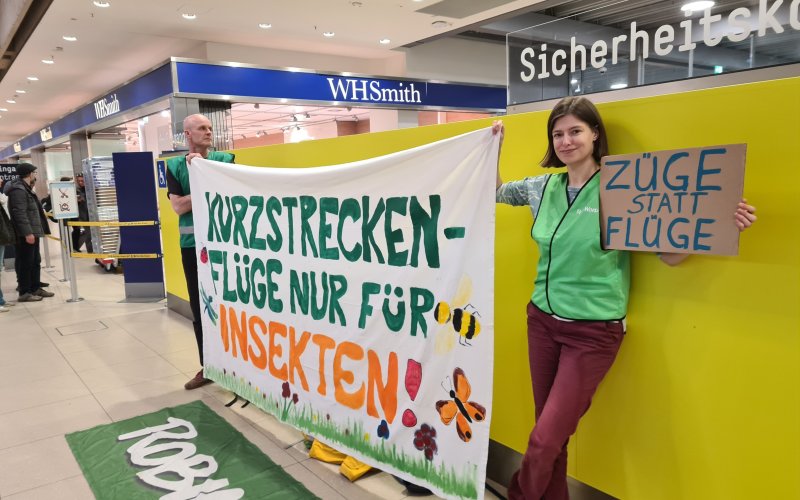 Aktive mit Protestbanner "Kurzstreckenflüge nur für Insekten" in der Abflughalle eines Flughafens