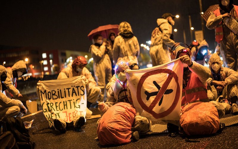 Foto von Menschen in weißen Anzügen die nachts auf der Straße sitzen. Auf den Bannern steht "Mobilitätsgerechtigkeit" und ein durgestrichenes Flugzeug