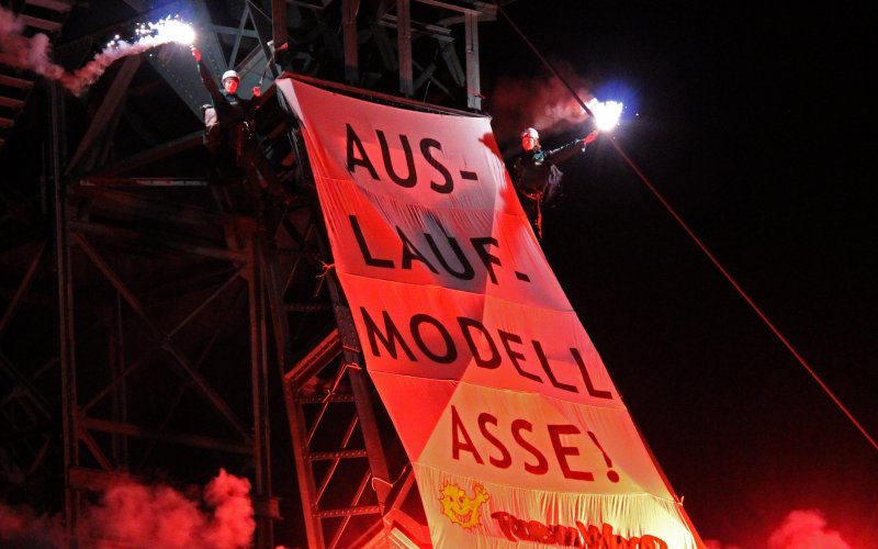 großes Banner am Förderturm der Asse. Die Aufschrift; Auslaufmodell Asse II - Rückholung sofort, Raus kommt es so oder so! -ROBIN WOOD. Es ist dunkel und die Aktivist*innen halten Pyros