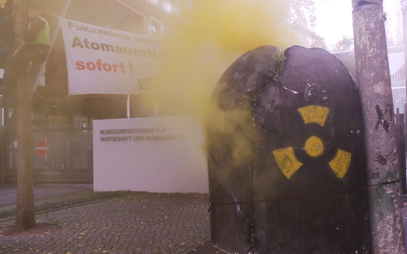 Ein AKW-Modell steht vor dem Ministerium für Wirtschaft und Klimaschutz, aus ihm kommt gelber Nebel, 2 Aktivisten von Robin Wood sind daneben Bäume geklettert mit einem Banner auf dem steht "Fukushima mahnt: Atomausstieg sofort", um gegen die Laufzeitverl