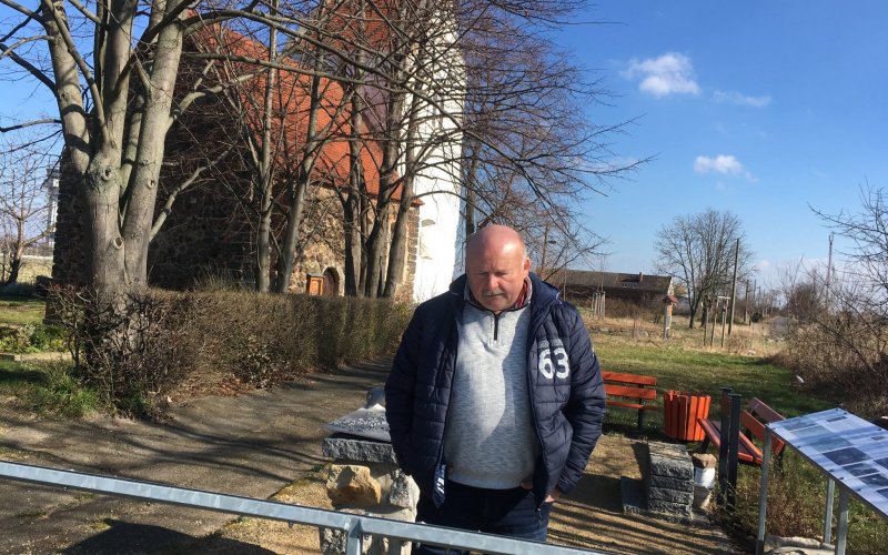Peter Richter steht vor einer Schautafel im Ort Kursdorf. Im Hintergrund ist eine verlassene Kirche zu sehen.