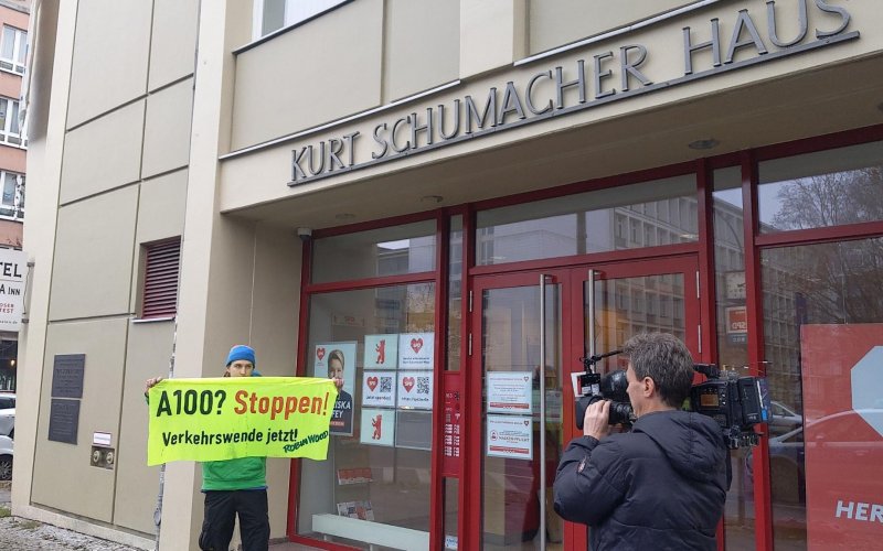 ROBIN WOOD Kameramann filmt Aktivisten, der ein Handbanner trägt. Aufschrift: "A100? Stoppen! Verkehrswende jetzt! ROBIN WOOD"