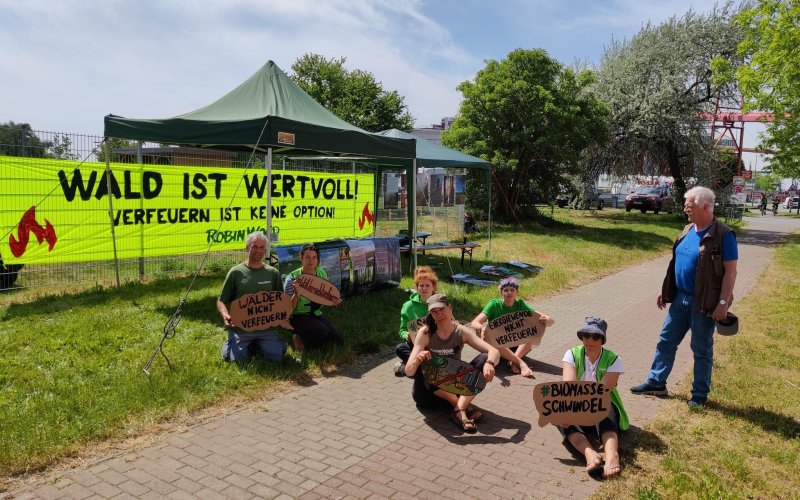 Aktive sitzen mit Protestschildern auf dem Boden vor einem Pelletwerk